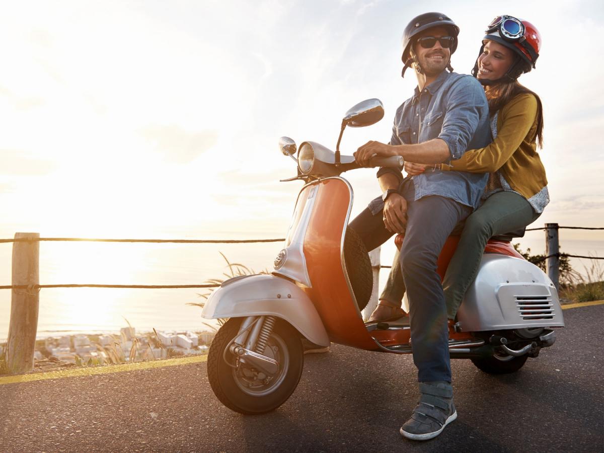 Ein Mann sitzt auf einem Roller und hinter ihm eine Frau, die sich an ihm festhält.